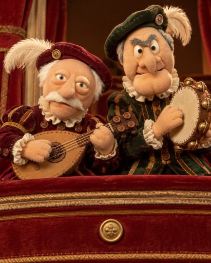 Two puppet-like characters in ornate historical costumes play a lute in a theater box.
